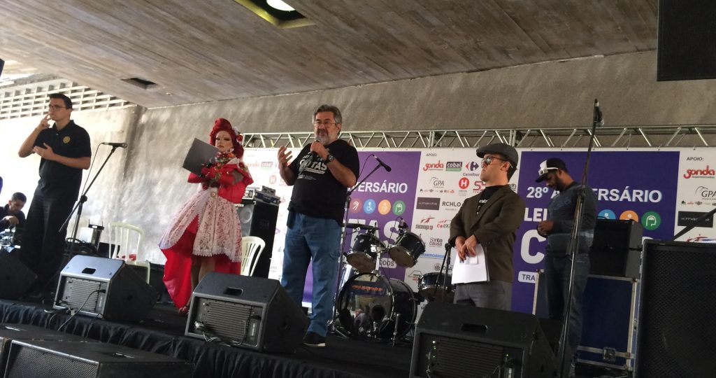 fotografia mostra o Dr Kal discursando ao lado dos mestres de cerimônia e do intérprete de Libras da festa do 28o. Aniversário da Lei de Cotas