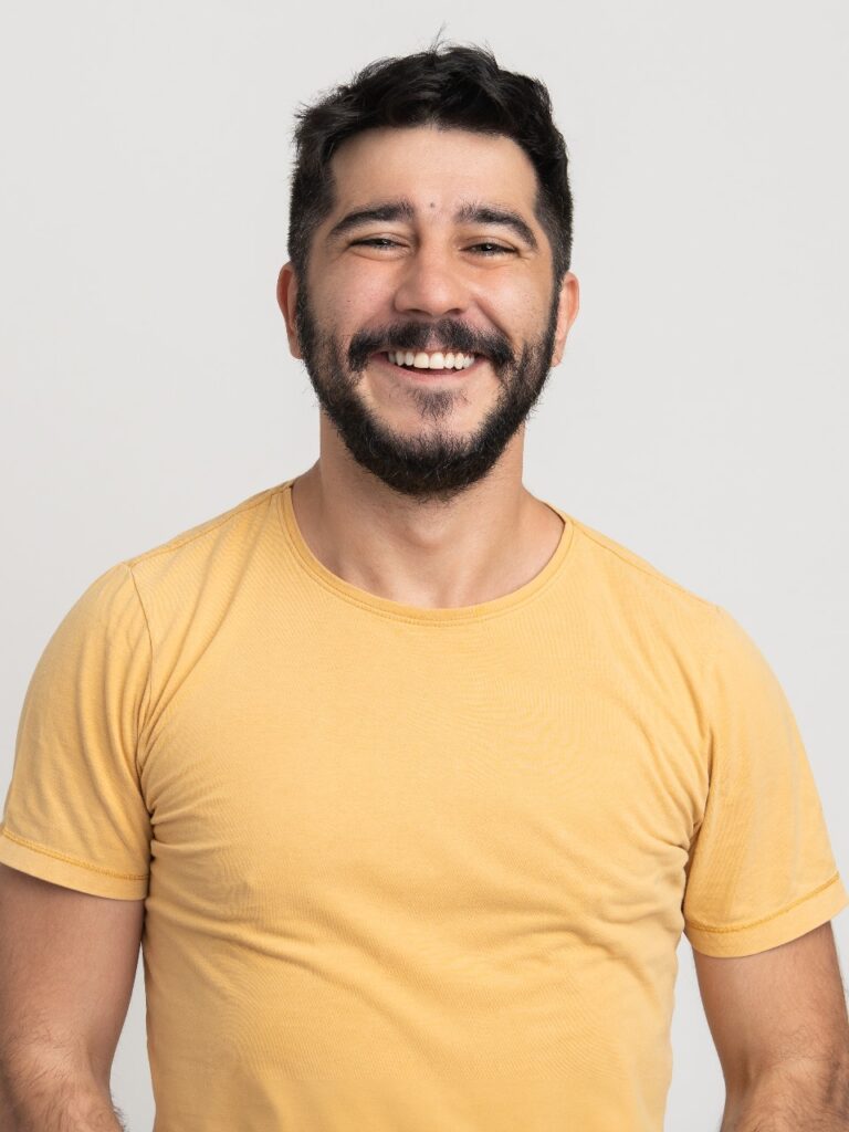 Wanderley é um homem de pele parda, cabelos pretos, barba e bigode pretos, olhos escuros, está sorrindo e usa uma camiseta lisa em tom alaranjado claro.