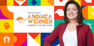 Andrea Werner é uma mulher branca, de cabelo e olhos castanhos, está usando camiseta branca e paletó vinho por cima, e está à frente de um banner com figuras coloridas gráficas, como seu nome e o texto "deputada estadual, gabinete da inclusão"