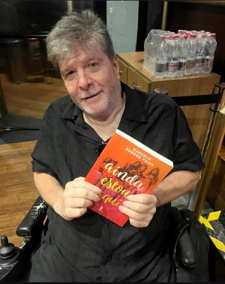 Foto do autor Marcelo Rubens Paiva, segurando o seu livro "Ainda estou aqui" com as mãos apoiadas no colo. Marcelo tem cabelo, bigode e barba prateados e bem aparados. É um homem branco de olhos castanhos. Usa camisa de manga curta e calças pretas. Está sentado em sua cadeira de rodas motorizada.